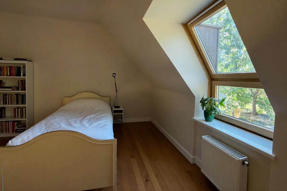 Zolderslaapkamer met bed, boekenkast en een dakkapelraam met uitzicht op de tuin